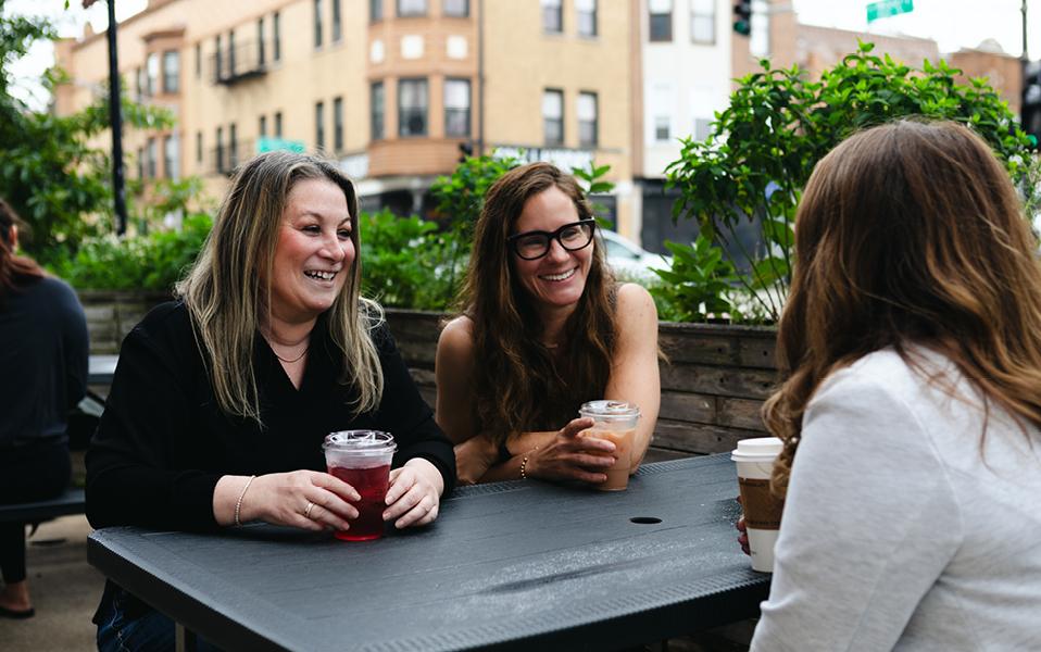 Three women talking at an outdoor café table with iced drinks